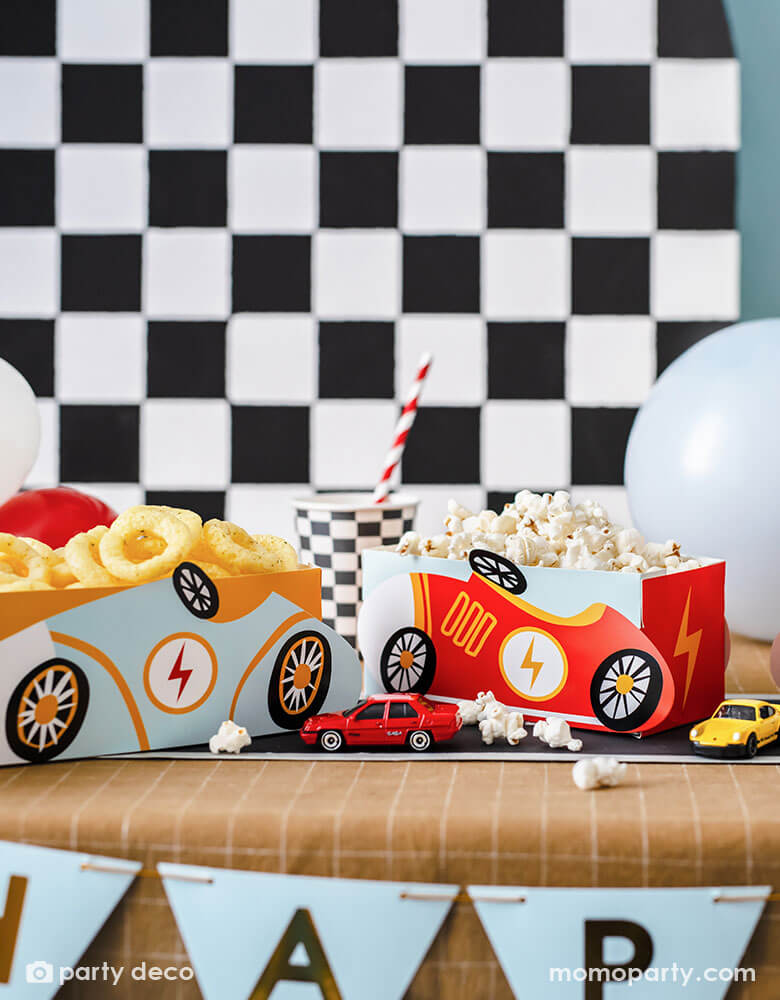 A modern kid's race car themed party table features Momopartys's race car snack boxes in light blue and red, filled with popcorn and onion ring chips. On the table there are checkered pattern party cups and race track table runner, with toy cars decorated. In the back there's a checkered backdrop decorated with some party balloons, making this a perfect inspo for kid's race car themed parties including a "Two Fast" second birthday party or a "Fast One" first birthday party."