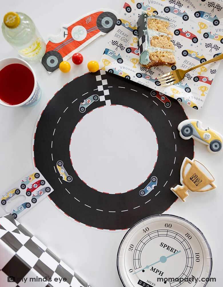 A festive kid's race car themed party table features Momopartys's race track placemats decorated with race car stickers. Around the placemats are race car pattern paper plates, napkins, party cups, speedometer shaped round plates and a checkered table runner by My Mind's Eye. On the table there are some matching race car sugar cookies and a slice of cake, making this a perfect inpo for a car themed bash for your child, be it a TWO FASt 2nd birthday, or a FAST ONE first birthday party.