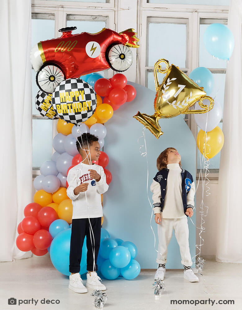 Two boys holding Momopartys's race car themed foil balloons including a red vintage race car shaped balloon, two checkered flag happy birthday foil balloons and a gold trophy shaped foil balloon, both kids standing in front of a pastel blue party backdrop wall which is decorated with colorful balloon garland with race car inspired colors including red, light blue, orange, gray and yellow. All makes this a festive and fun kid's birthday celebration.