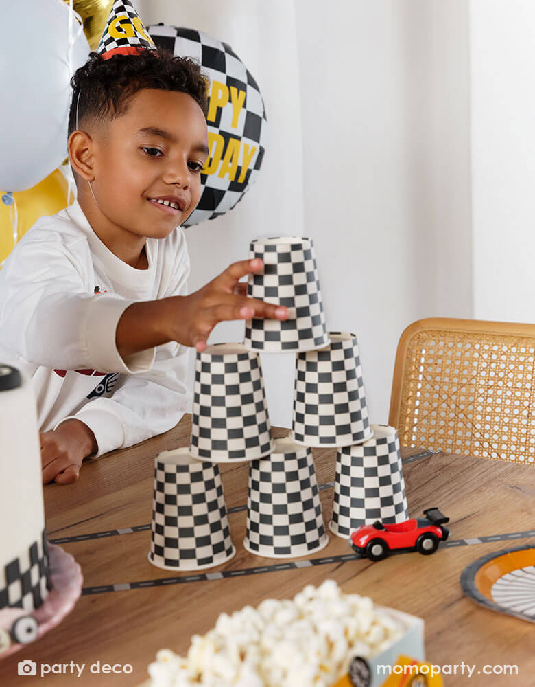A boy wearing Momopartys's checkered race car party hat stacking the checkered flag party cups to make a cup tower on a party table, next to the cups are some party supplies from Momopartys including wheel shaped plates, race car track tape, and some toy cars.
