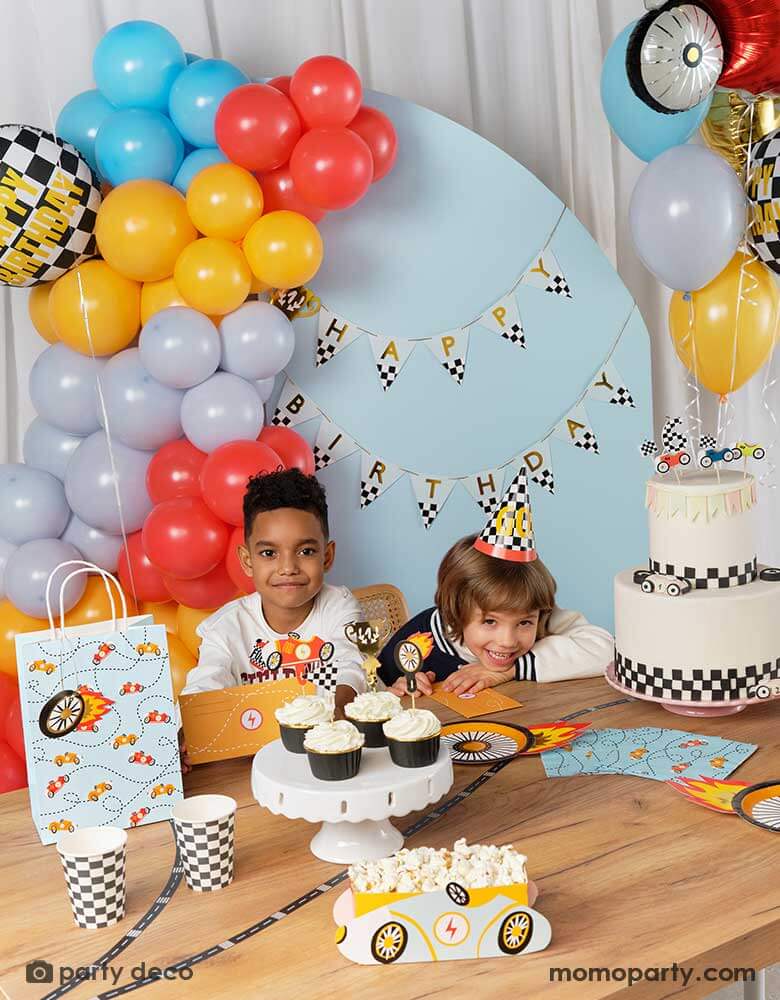 Two boys in a race car themed birthday party with them sitting at a table filled with race car themed party supplies from Momopartys including wheel shaped party plates, checkered party cups, race car themed napkins and party bags. With birthday cake and cupcakes topped with race car candles and cupcake toppers. Behind them is a blue backdrop wall decorated with trophy happy birthday banner from Momopartys and colorful balloon garland in the race car themed colors.