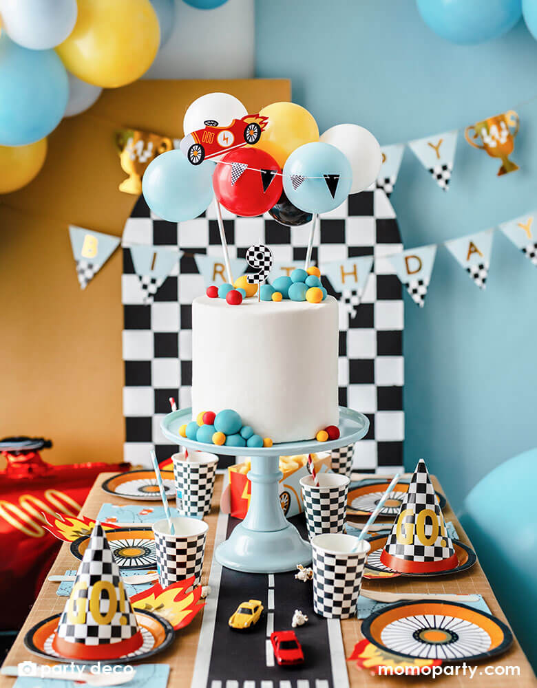 A kid's race car themed party table filled with race car themed party supplies from Momopartys including wheel shaped plates, checkered flag GO! party hats, blue race car large napkin, and checkered party cups. On the table there is a modern white birthday cake topped with Party Deco's race car balloon topper. In the back you can see a checkered backdrop with Momopartys's race car trophy happy birthday banner along with some colorful balloon garland hung on it, perfect for kid's Two Fast or Fast One party.