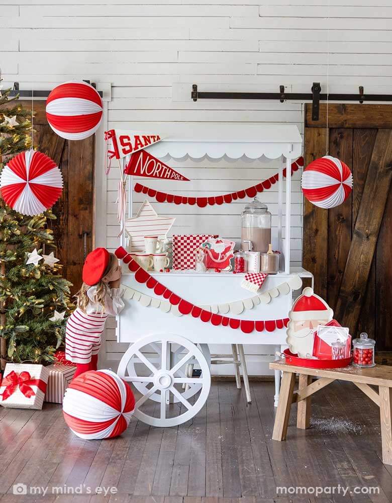 A girl in an adorable Santa themed hot cocoa station set up for a kid's Holiday party featuring a white dessert cart with hot chocolate and Momopartys's Dear Santa party supplies including Santa shaped party cups, checkered plates, napkins, Santa on a sleigh shaped plates and Dear Santa envelope shaped plates. Around the hot chocolate station are two felt party pennant with Santa & Northpole written on them, red scalloped shaped party garlands and some red and white honeycomb balls hung from the ceiling.
