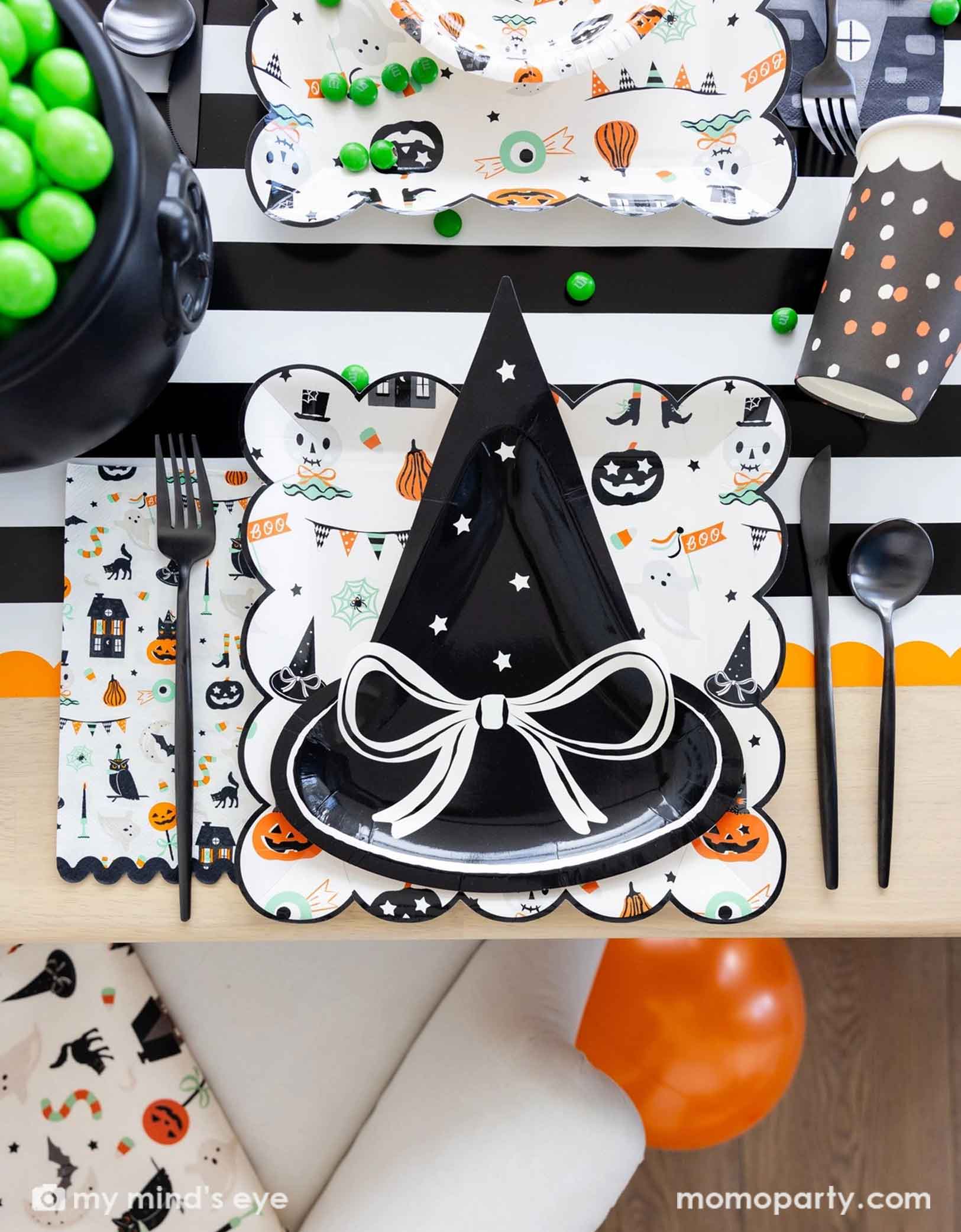 A overhead shot of a festive Halloween-themed table setting with the witch hat shaped plate, Trick or Treat dinner plates and bowls, the haunted mansion shaped napkins and black cutlery set on a black orange and white striped table runner fro Momopartys. On the table there's a cauldron filled with green gum balls and some green candies spread across the table. A perfect set up for kid's friendly Halloween celebration.