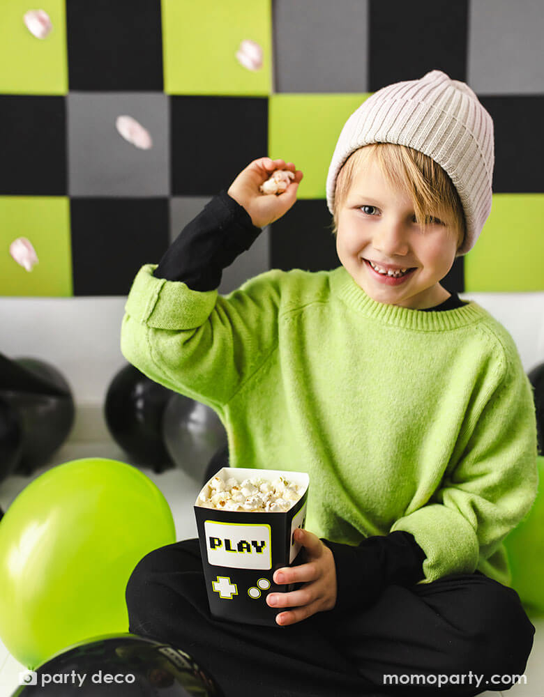 A boy in the classic video game black and green outfit sitting on the floor surrounded by black and green party balloons. He's holding Momopartys's 2.75 x 2.75 x 4.75 inches black video game snack box by Party Deco, filled with popcorn, trying to throw the popcorn at his friend.