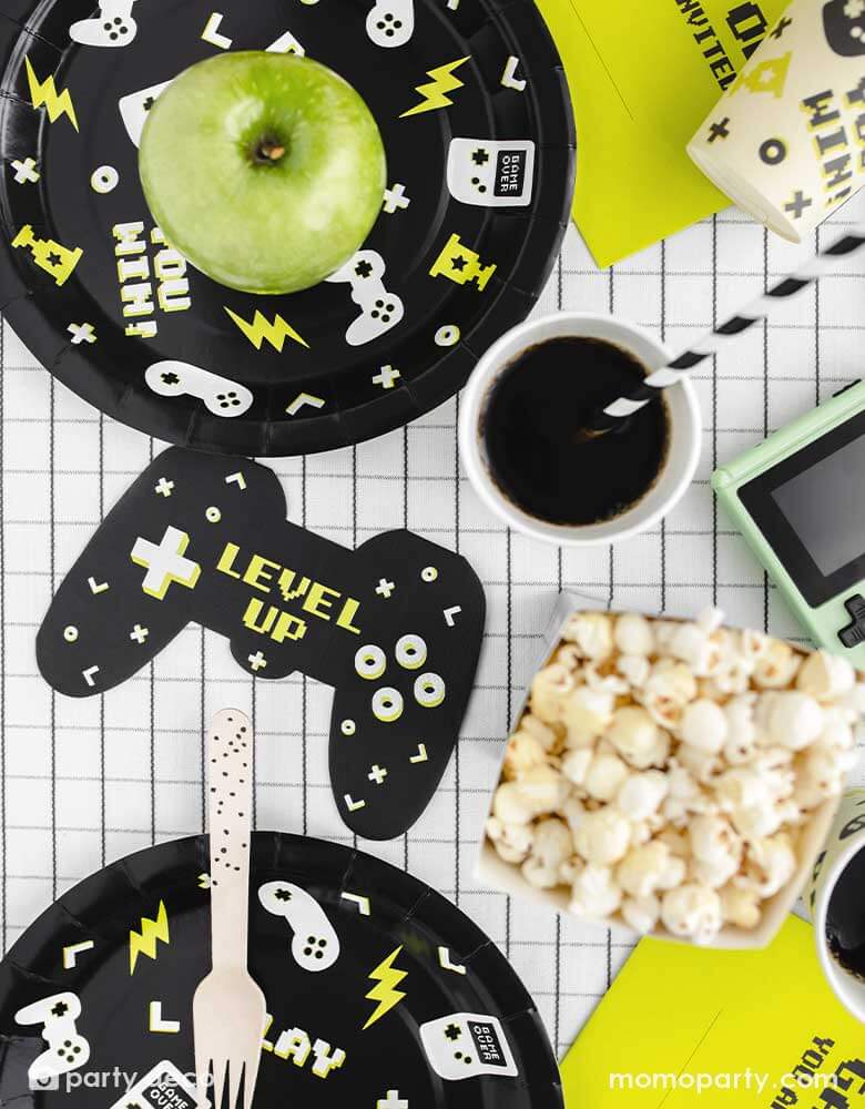 A video game themed party table features Momopartys's black Video Game round party plates with video game devices and controller motifs on them. Next to the plates are a game controller shaped napkins with "LEVEL UP" message on them, with the coordinating video game party cups with black striped paper straws from Momopartys. On the table covered with black and white grid tablecloth, the are some popcorns and an green apple as snacks, ready for a fun game time!