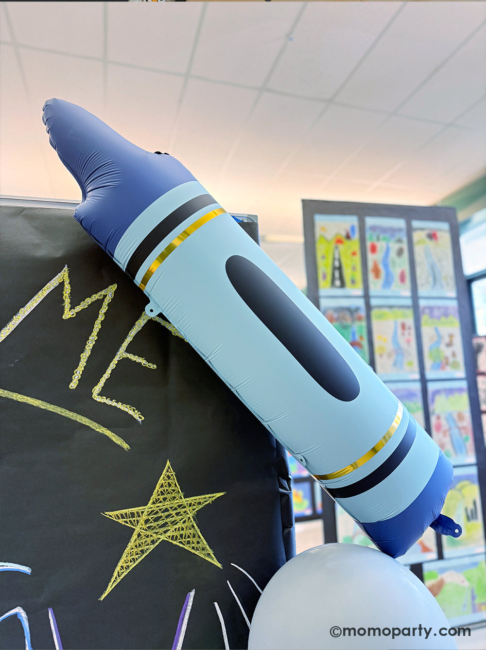 A close up shot of Momopartys's 40" x 9" blue crayon shaped foil balloon decoration next to a chalk board at a kid's art show in a school event. Its bright color and fun design is perfect for kid's school themed celebration, be it a back to school party, a first day of school celebration or any kids event at school.