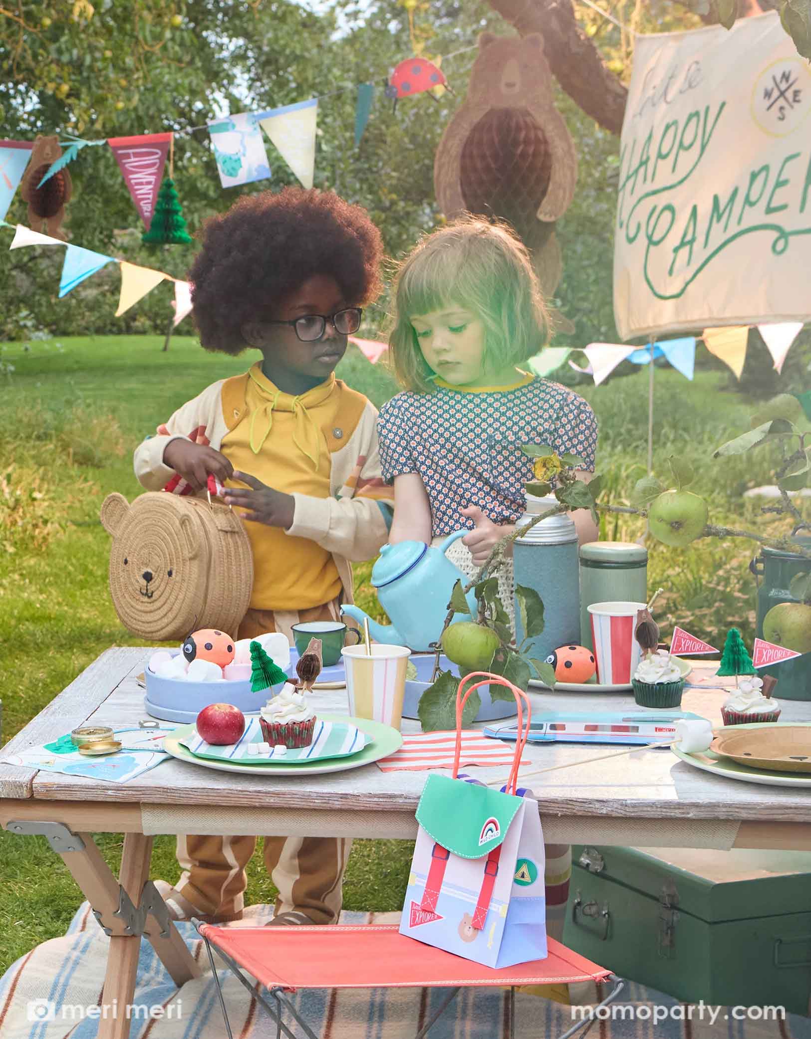 Two cute school aged girls standing next to a camping themed party table featuring multiple Meri Meri camping themed party supplies from Momopartys, including camping RV shaped plates, bear plates, compass shaped napkins, backpack shaped party bags and ladybug surprise balls. Above them hung a 10ft camping icon party garland featuring camping themed pennants and bear honeycomb. With a sign says "Happy Camper" this makes a great inspo for kid's camping themed party decorations.