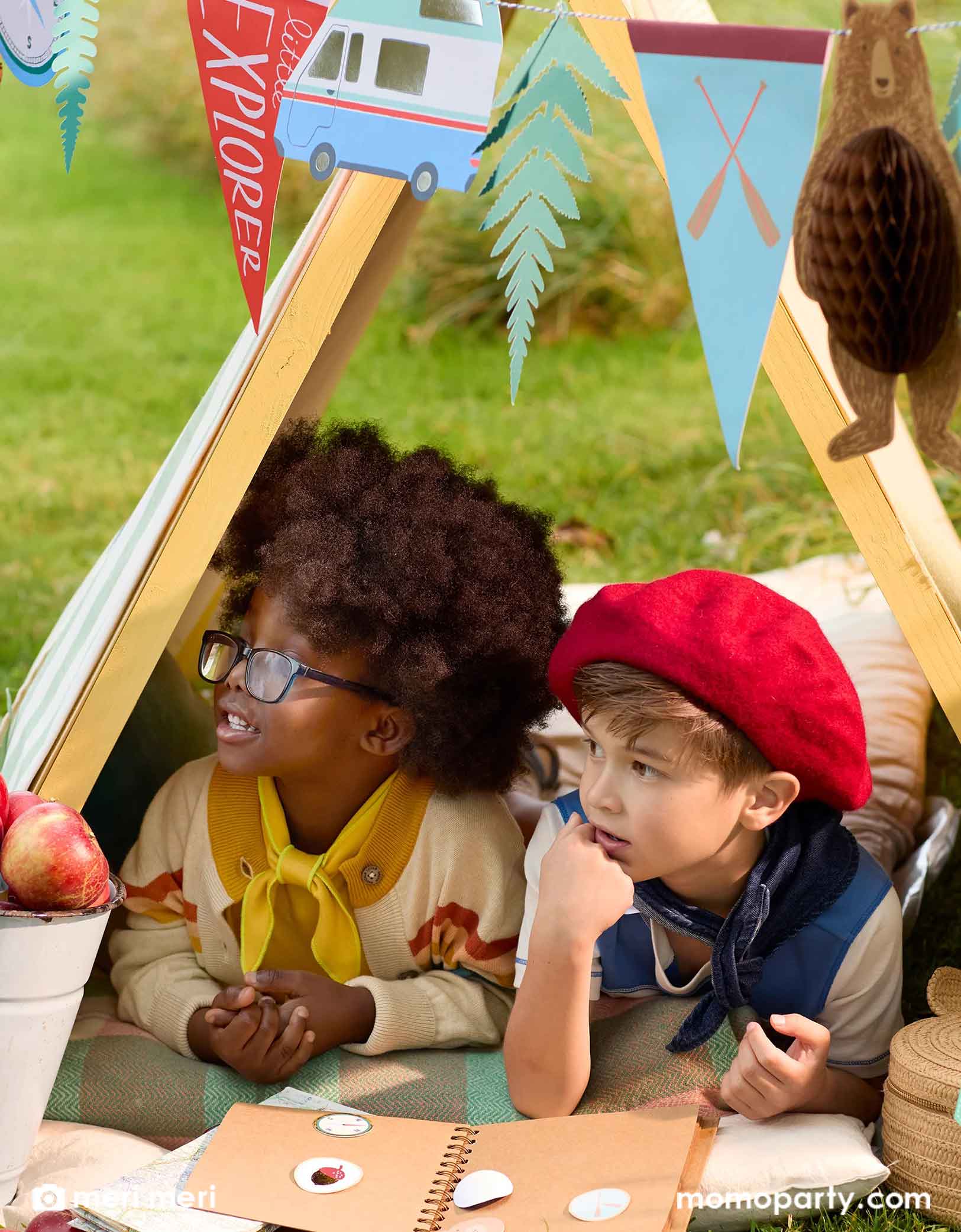 Two cute school aged kids dressed like boy scouts lying under a camping tent decorated with Momopartys's 10 ft adventure camping icon party garland featuring camping themed party pennants and bear honeycomb. Around the kids are some apples and a book of camping stickers collection.