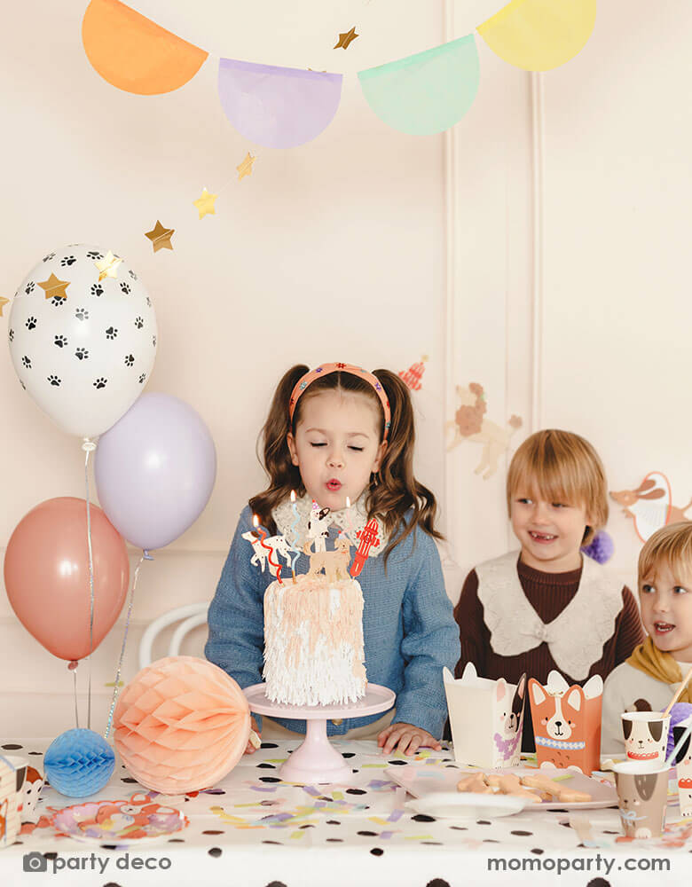A girl blows birthday candles form a dog themed birthday cake topped with Momopartys's puppy dog cake toppers in a puppy dog themed birthday party. This party features various dog themed party supplies from Momopartys including party banners, paw balloons, corgi standing foil balloon, Dalmatian print dots tablecloth, dog shaped snack boxes, party cups and honeycomb decorations in soft pastel colors. These cute dog themed party goods make a festive touch to kid's birthday bash!