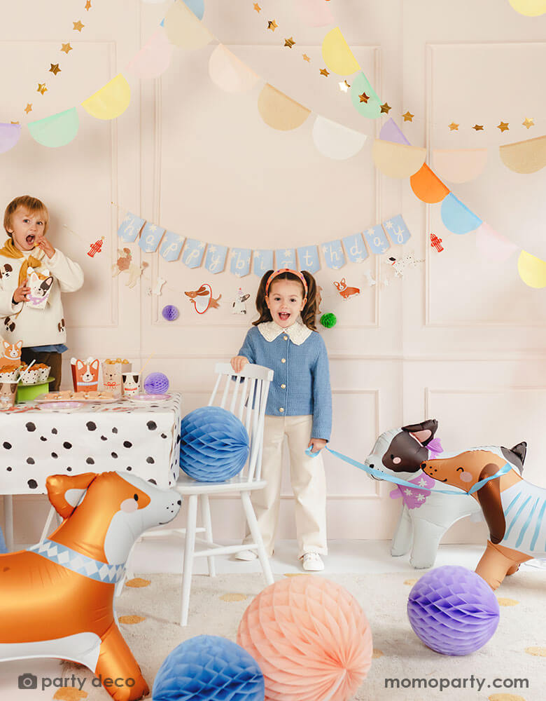 A kid's puppy dog themed birthday party set up featuring various dog themed party supplies from Momopartys including party banners, paw balloons, corgi, bull dog and Dachshund standing foil balloons, Dalmatian print dots tablecloth, dog shaped snack boxes, party cups and honeycomb decorations in soft pastel colors. These cute dog themed party goods make a festive touch to a kid's birthday bash!