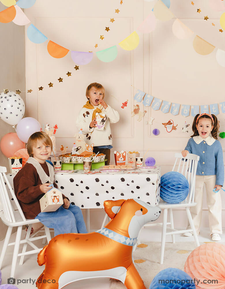 A kid's puppy dog themed birthday party set up featuring various dog themed party supplies from Momopartys including party banners, paw balloons, corgi standing foil balloon, Dalmatian print dots tablecloth, puppy dog shaped snack boxes, party cups and honeycomb decorations in soft pastel colors. These cute dog themed party goods make a festive touch to kid's birthday bash!