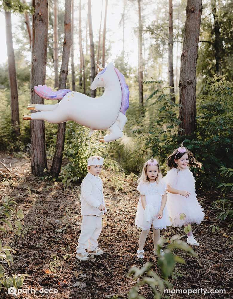 Three kids dressed in white clothing with party hats in an enchanting forest with Momopartys's enchanting unicorn shaped foil balloon floating in the air, making this a dreamy scene for kid's special celebration.