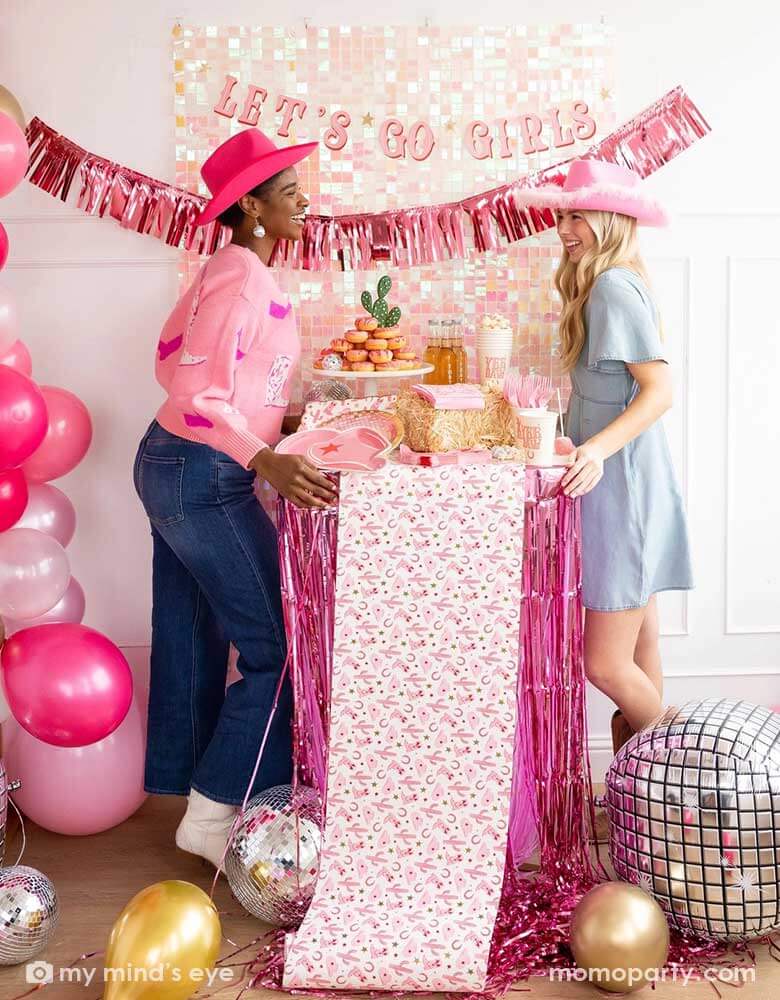 Two young ladies in their Western rodeo cowgirl inspired outfits and pink cowgirl hats in a disco cowgirl themed bachelorette party decorated with cowgirl themed party supplies, decorations and balloons from Momopartys. They're facing each other by a table decorated with cowgirl party supplies including plates, napkins and a paper table runner. There's a donut cake, beverages and various treats on the table. Behind them is a iridescent wall adorned with "Let's Go Girls" fringe party banner from Momopartys.