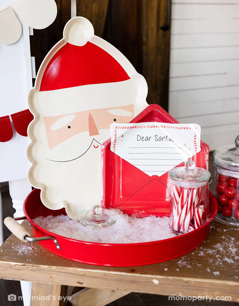 A hot cocoa station set up ideas in a kid's Holiday party featuring Momopartys's Letter to Santa envelope shaped plate, Santa shaped bamboo tray along with jars of candy canes, red gum balls in a red round tin tray. With the artificial snow decoration around, this make s a whimsical decoration for a festive Christmas celebration!