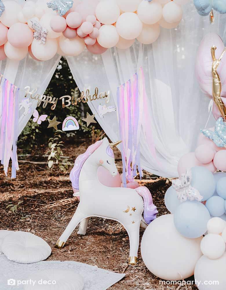 An enchanting unicorn themed birthday party backdrop decoration featuring Momopartys's 37.5" x 34" unicorn standing foil balloon around beautiful pastel colored balloon garlands adorned with butterfly foil balloons and a gold Happy Birthday banner with pastel streamers in the front of a dreamy curtain, making this a whimsical scene for a girly's whimsical unicorn themed birthday party.