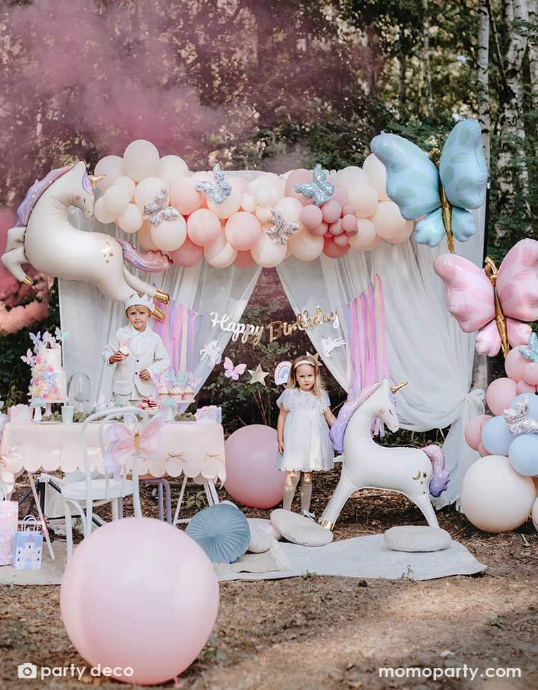 Two kids dressed up as fairies standing in an enchanting unicorn themed birthday party backdrop decoration featuring Momopartys's 37.5" x 34" unicorn standing foil balloon around beautiful pastel colored balloon garlands adorned with butterfly foil balloons and a gold Happy Birthday banner with pastel streamers in the front of a dreamy curtain. In the front there's a party table with a two tier birthday cake and cupcakes, making this a whimsical scene for a girly's whimsical unicorn themed birthday party.