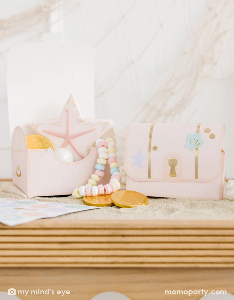 Two under the sea treasure chest box shaped treat boxes from Momopartys on a wooden party table. These light pink treat boxes are decorated with adorable pastel colored seashell and sea star stickers. In the treat boxes there's a pink sea star shaped sugar cookie, gold chocolate coins and a candy necklace, making this a great party favor idea for a kid's mermaid or under the sea themed birthday party.