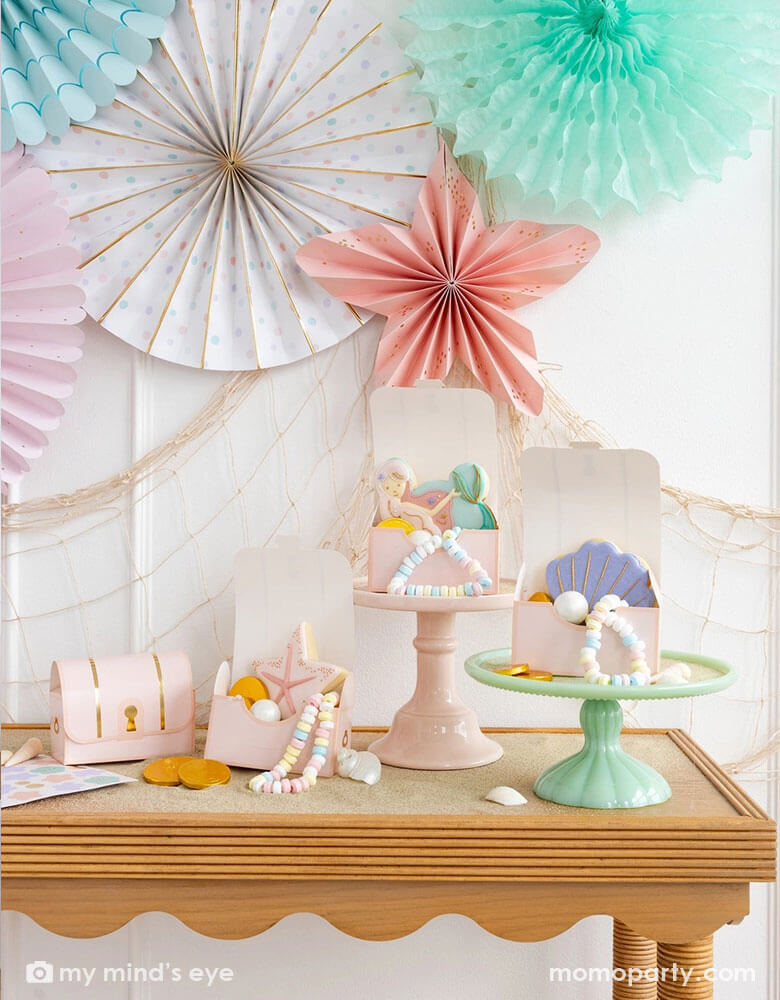 A mermaid party table with Momopartys's pink under the sea treasure chest treat boxes by My Mind's Eye. In these treat boxes are some pastel colored treats, mermaid themed sugar cookies with designs of a mermaid, a seashell and a sea star in soft pastel colors. Some of them are placed on pastel colored cake stands. The wall behind is decorated with Momopartys's under the sea paper fan set in pastel colors of aqua, pink, lilac, teal, cream and peach, with a fishing next hung underneath.