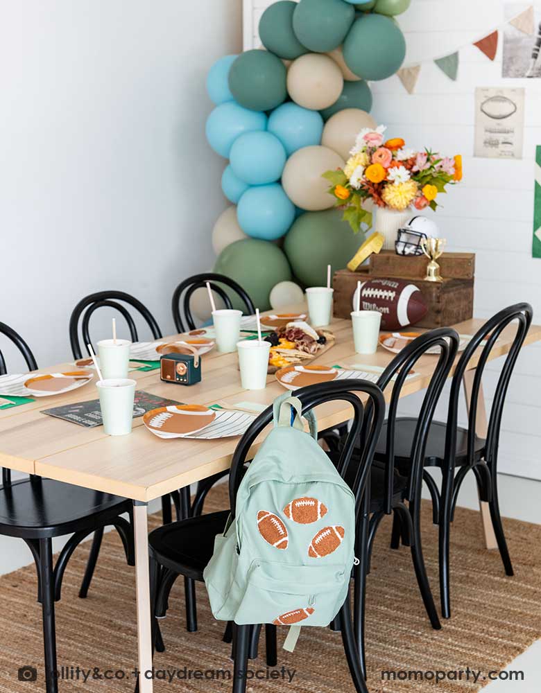 A kid's football birthday party set up with a modern party table filled with football themed tableware from Momopartys. In the center of the table there's a cheese charcuterie board and some decorations like a football, a trophy and a football helmet as the centerpiece. On one of the chairs, there's a backpack with football patches on it. On the wall there are some old school football party banners, posters and party balloons to add some festivities to the celebration!