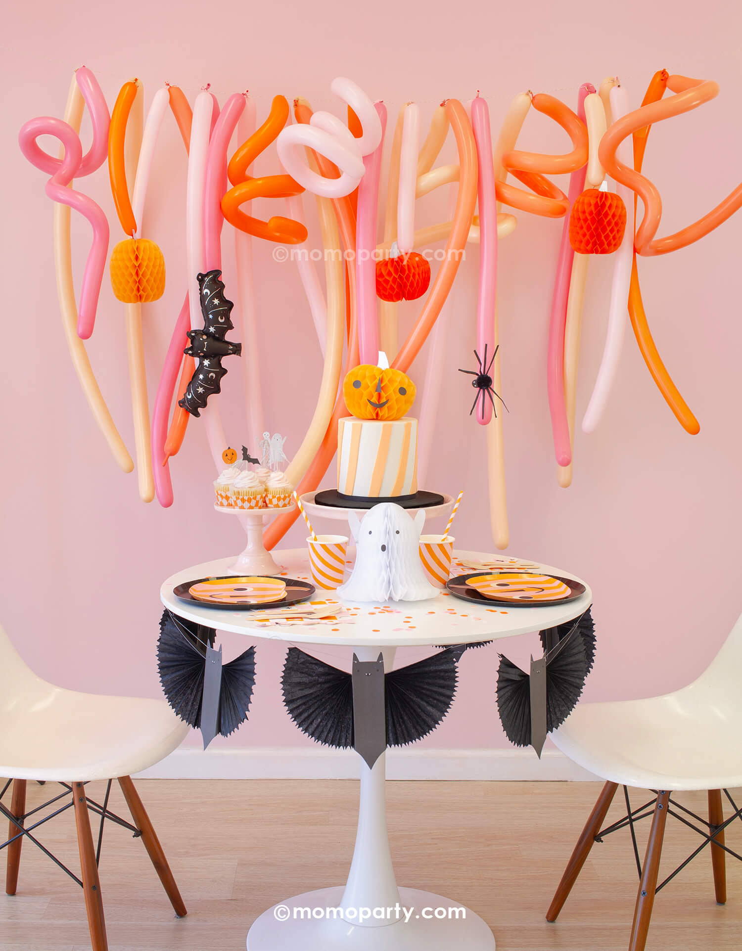 Momo Party's Trick or Treat Pink Halloween box set up featuring a festive long balloon backdrop with curling long balloons in coral, pink, rose and peach colors. On the balloon backdrop it's decorated with paper pumpkin honeycombs and a mini bat foil balloon. In front of the balloons, a small round table is filled with Halloween party supplies including pumpkin shaped plates, a bat garland, ghost and pumpkin honeycomb decorations from momopartys.shop.