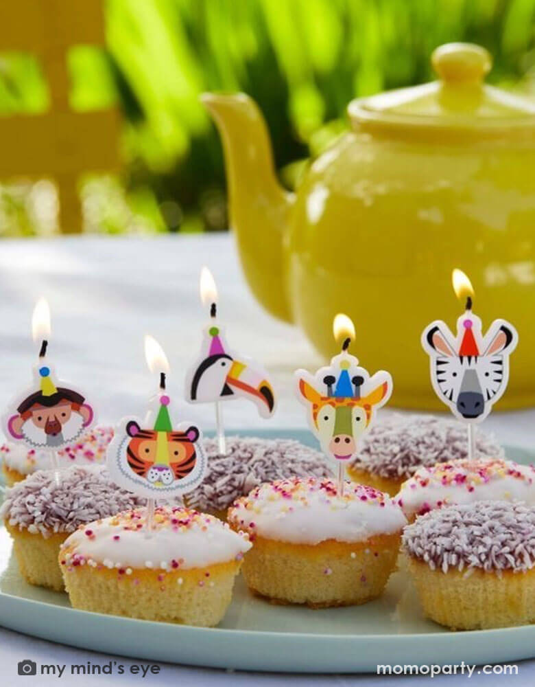 A few birthday cupcakes topped with Momopartys's animal birthday candles of a tiger, a zebra, a monkey, a toucan and a giraffe by Talking Tables. In the back is a yellow tea pot on the table ready for an afternoon time party.