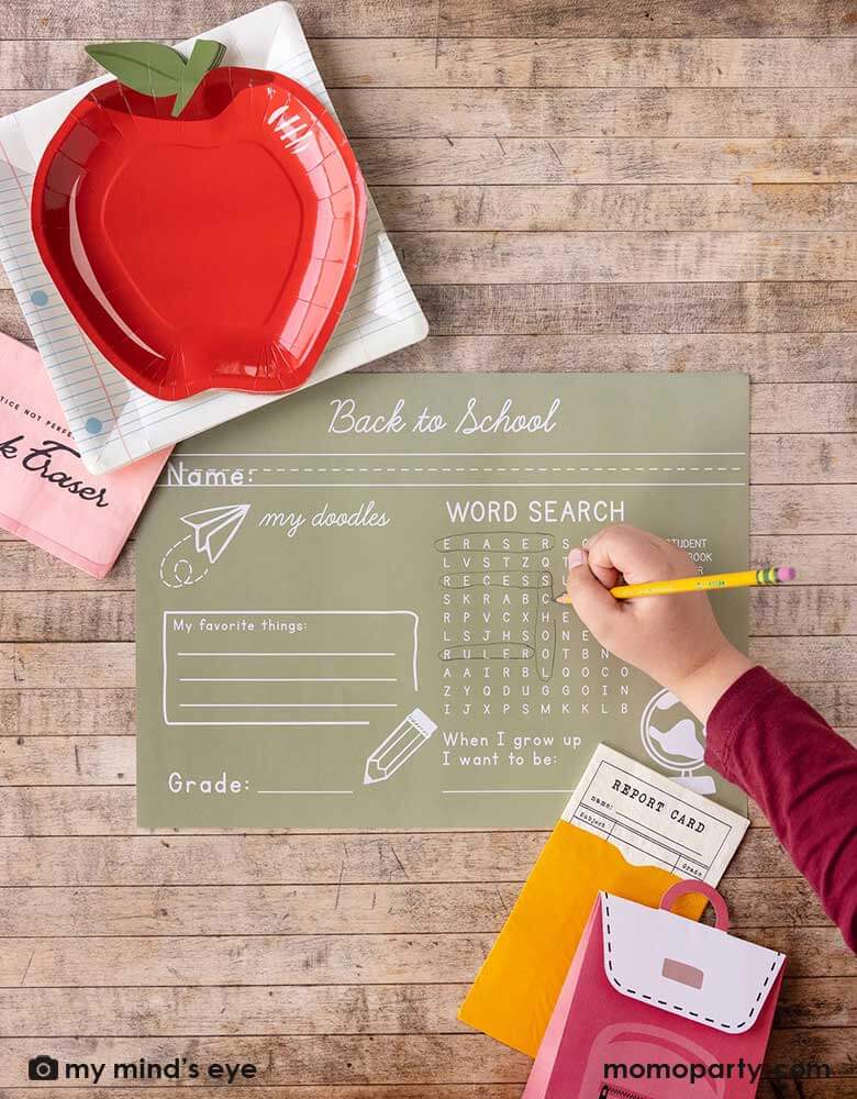 A kid holding a pencil playing work search game on Momopartys's back to school paper tablemat by My Mind's Eye. Around the tablemat, there are apple shaped plates layered with notebook square plates and pink eraser shaped napkins, at the right bottom corner of the photo, there are a report card shaped napkin with backpack shaped treat box. All in all makes a great inspiration for kid's back to school celebration.