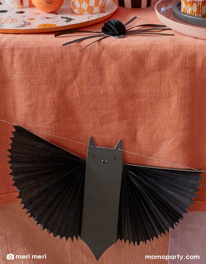 A pink Halloween party table decorated with Momopartys's Halloween bat garland by Meri Meri. In the back you can see a corner of a table with honeycomb spider decoration and a variety of Halloween themed tableware including paper dinner plates, cups and cupcake toppers with Halloween characters including pumpkins, bats, ghosts and more.