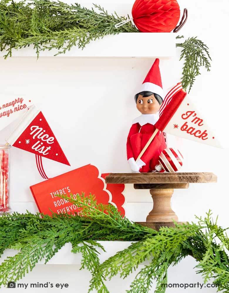 An elf on the shelf sitting on a white shelf decorated with green Christmas tree branches and leaves, on his hands held a red and white Momopartys's Christmas Elves on the Shelf felt pennant banners flag by My Mind's Eye featuring words "I'm Back.", along with other felt banner flags featuring "Never Naughty Always Nice", along with some red jelly beans in a candy jar and a red honeycomb ball decoration. All makes a great idea for kid's Holiday elf on the shelf Christmas home activity.
