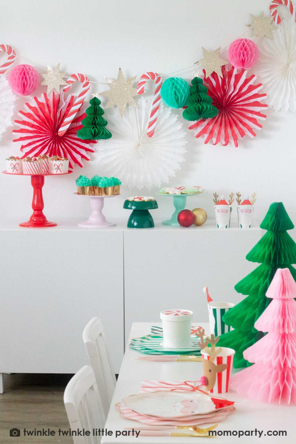 A Christmas party table featuring Momopartys's festive striped party tableware in pink red and green with Christmas tree honeycombs as the centerpiece. In the back is a Christmas dessert table decorated with Momopartys's Christmas honeycomb decorations featuring candy canes, honeycomb Christmas trees and mint honeycomb balls. On the table there are multiple cake stands in red, pink, green and mint with Holiday treats like cookies, cupcakes and baked food, making this a perfect Holiday party inspo.