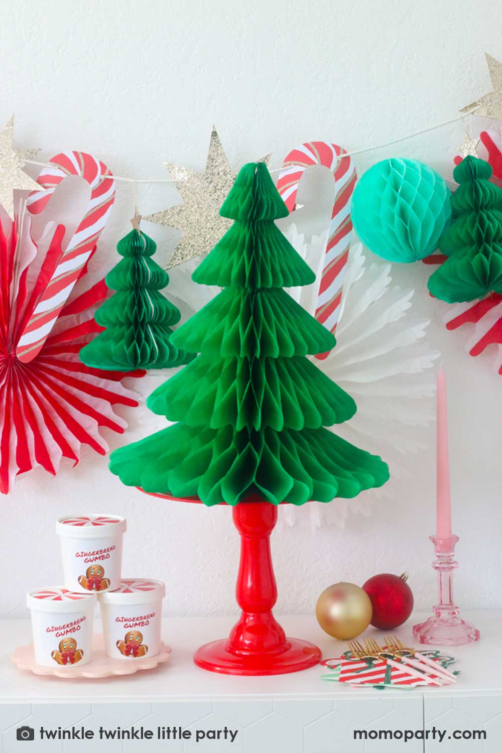 A Christmas Dessert Table decorated with Momopartys's Christmas honeycomb decorations featuring candy canes, honeycomb Christmas trees and mint honeycomb balls. On the table there's a red cake stand with Momopartys's 17" light green honeycomb Christmas tree on it, a pink candle on a clear pink candle stand next to Meri Meri's present with a bow shaped napkins in festive colors of red and green. On the left, there are some peppermint treat boxes stacked, making this a perfect Holiday party inspo.