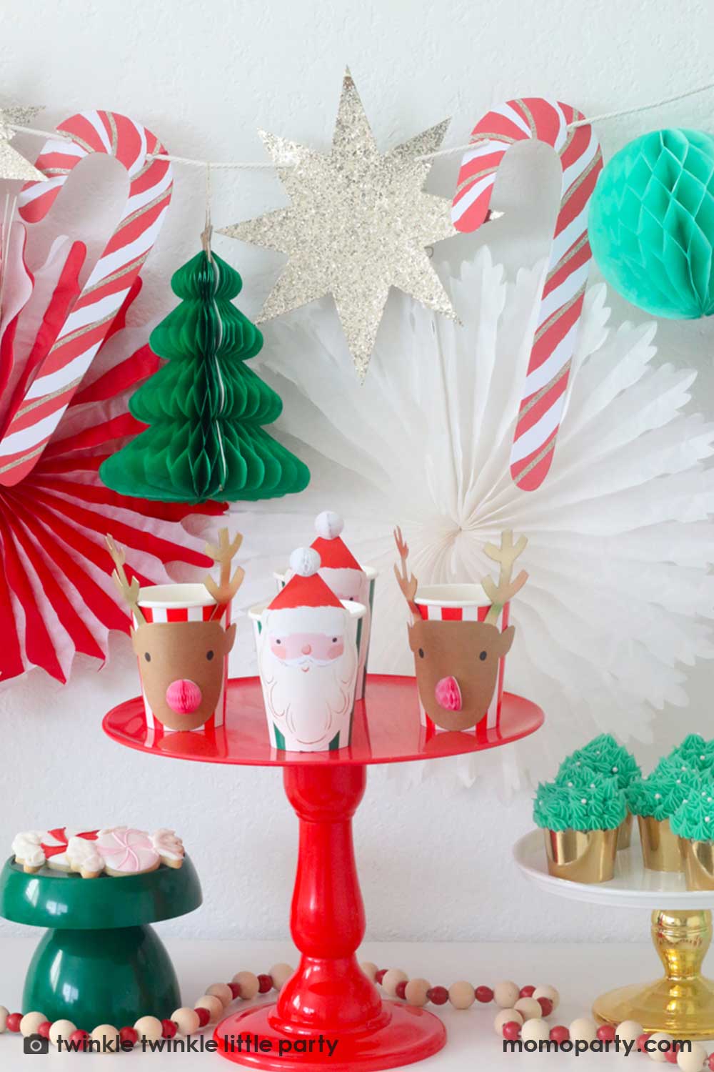 A Christmas Dessert Table decorated with Momopartys's Christmas honeycomb decorations featuring candy canes, honeycomb Christmas trees and mint honeycomb balls. On the table there's a red cake stand with Meri Meri's Christmas honeycomb party cups in the design of Santa and reindeer, and a gold cake stand with Christmas tree buttercream cupcakes and a small dark green cake stand with peppermint sugar cookies, making this a perfect inspo for a festive Holiday party setup.