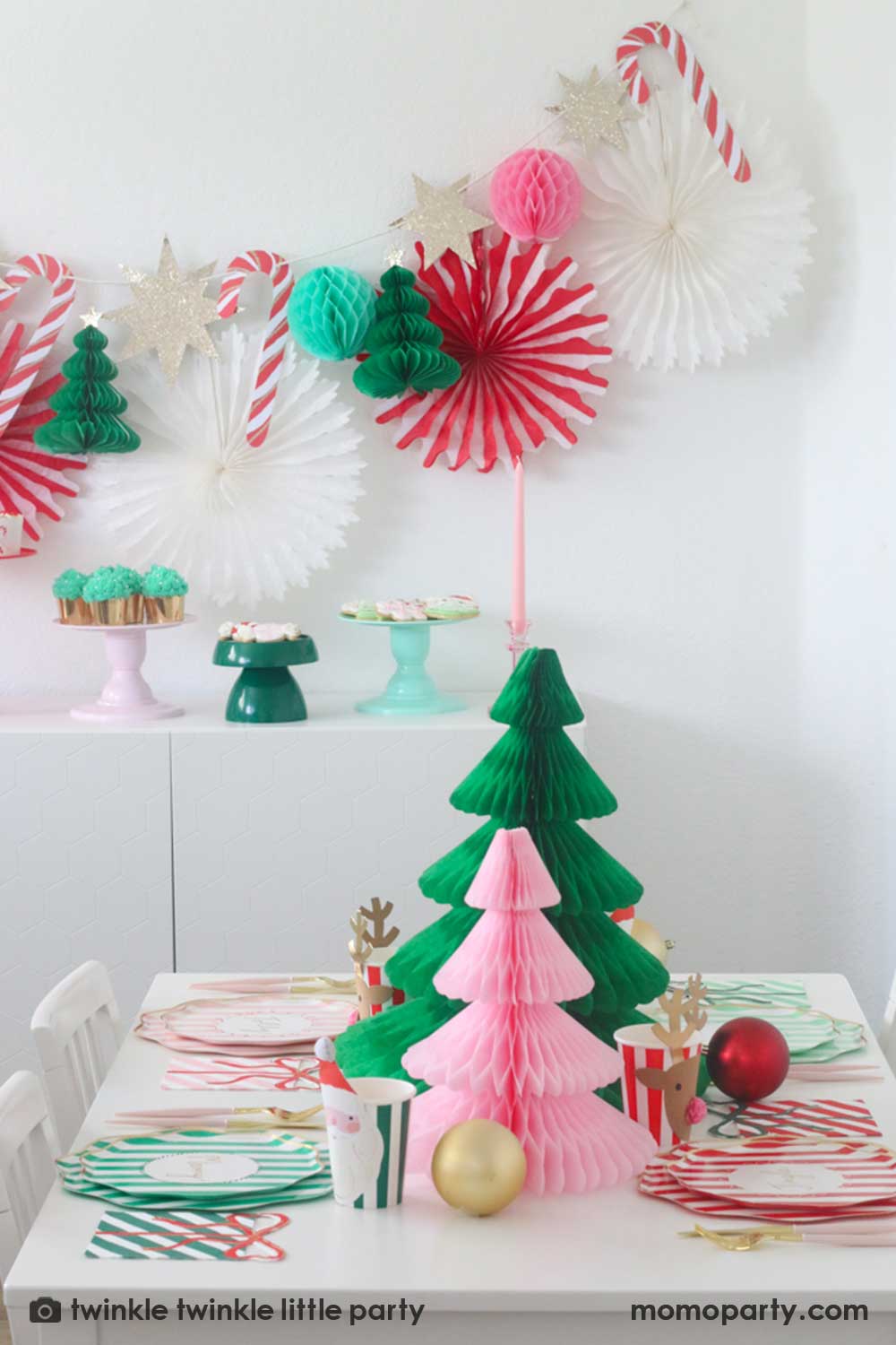 A Christmas party table featuring Momopartys's festive striped party tableware in pink red and green with Christmas tree honeycombs as the centerpiece. In the back is a Christmas dessert table decorated with Momopartys's Christmas honeycomb decorations featuring candy canes, honeycomb Christmas trees and mint honeycomb balls. On the table there are multiple cake stands in red, pink, green and mint with Holiday treats like cookies, cupcakes and baked food, making this a perfect Holiday party inspo.