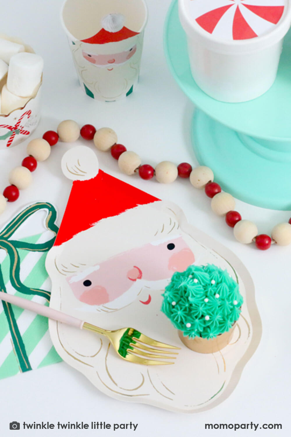 A festive Christmas tablescape featuring Momopartys's Jolly Santa Plate, present with a bow shaped napkin and Christmas honeycomb party cup in Santa design by Meri Meri. On the Jolly Santa plate there is a buttercream Christmas tree cupcake in festive gold foil baking cups and a festive red and white pom pom banner around the tablescape, making this a modern Holiday placeset inspo for a festive Holiday party.