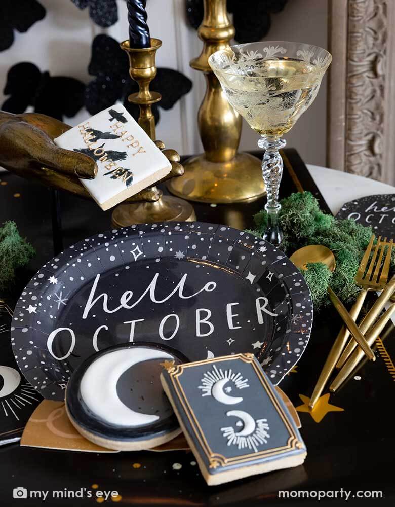 A mystical Halloween table decorated with Momopartys's moons and stars themed party supplies including a crystal ball shaped plate, moons and stars gold foil napkins, moons and stars table runner by My Mind's Eye. With gold metal candle stands holding black candles and vintage gold utensils with Halloween themed sugar cookies and black glittered butterfly decorations on the wall, all makes a perfect inspo for an elegant and chic wizard and witch themed Halloween party decorating idea.