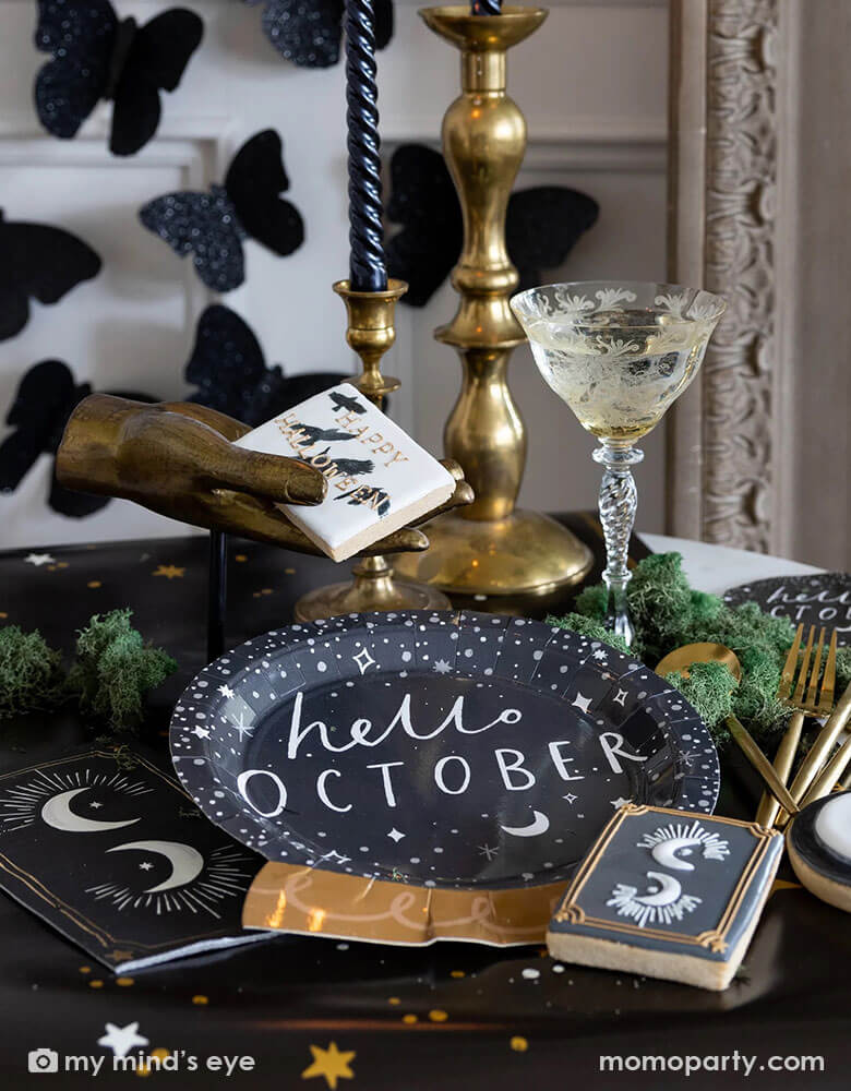 A mystical Halloween table decorated with Momopartys's moons and stars themed party supplies including a crystal ball shaped plate, moons and stars gold foil napkins, moons and stars table runner by My Mind's Eye. With gold metal candle stands holding black candles and vintage gold utensils with Halloween themed sugar cookies and black glittered butterfly decorations on the wall, all makes a perfect inspo for an elegant and chic wizard and witch themed Halloween party decorating idea.