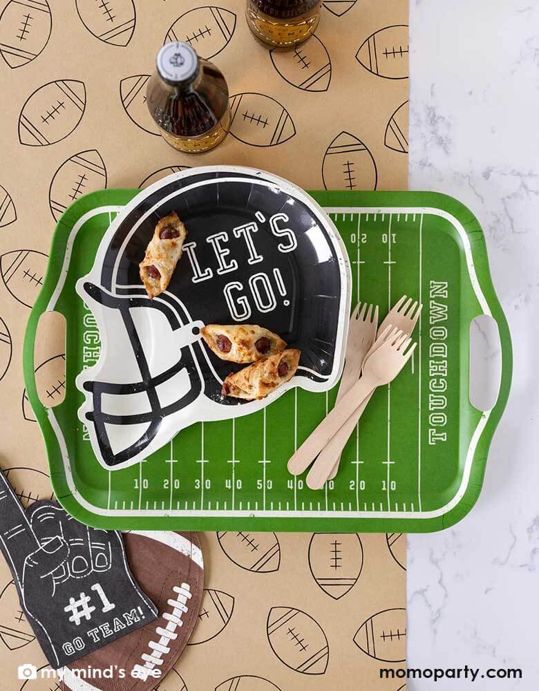 A football party table features Momopartys's Football Field bamboo reusable tray, on the tray it's a black helmet shaped plate featuring "Let's Go"" on it, no the plates there some party snacks and around the tray there are Momopartys's football shaped napkins and #1 finger shaped black napkin on a light brown table runner with football illustration on it. With party drinks around, it makes a great party table decoration for a fun football season watch party!