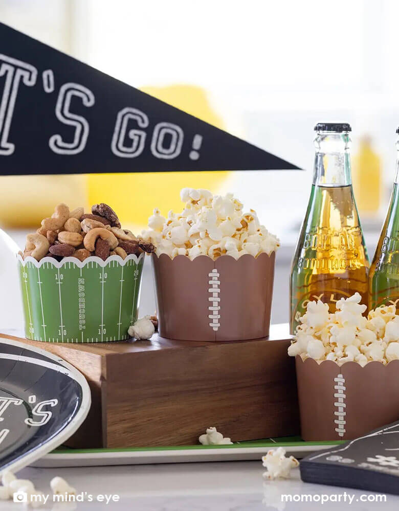 A football themed party table featuring Momopartys's jumbo football food cups filled with watching party snacks including popcorn and mixed nuts. Next to them is some root beer bottles and "Let's Go" felt party pennant by My Mind's Eye.