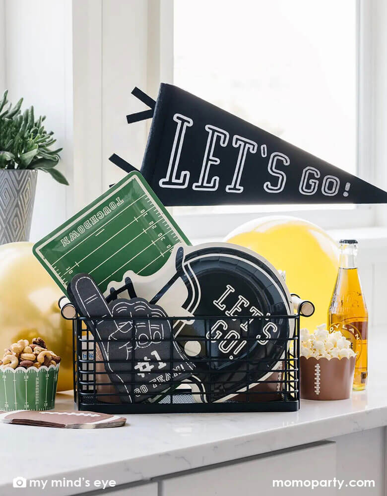 An occasion basket filled with Momopartys's football themed party decorations including a "Let's Go" felt party pennant, football field shaped serving plates, black football helmet shaped plates, black #1 finger shaped napkins and football themed foodcups filled with watching party snacks including popcorn and nuts, next to it are some yellow party balloons and some root beer bottles. Perfect for a watching game, super bowl party in football season!