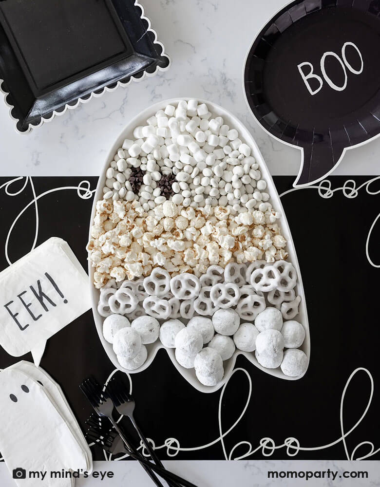 A black and white Halloween table featuring Momopartys's 12" x 16" ghost shaped bamboo reusable tray, filled with all sorts of Halloween treats and goodies including white donut holes, white sugar coated pretzels, popcorn, white chocolates and marsh mellow. Around it it's Momopartys's black boo shaped plates, ghost shaped napkins with EEK! shaped napkins on a black table runner with "boo" scripted font written over it. All makes a cool and spooky ideas for a kid's Halloween party table setting inspiration.