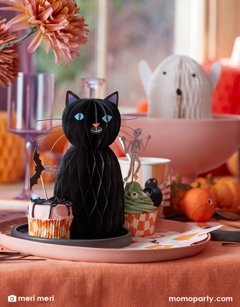 A pink and orange Halloween party table decorated with Momopartys's Halloween black cat and ghost honeycomb decorations by Meri Meri and a variety of Halloween themed tableware and cupcake toppers in the design of bats, skeletons, pumpkins and spiders. Makes it a whimsical inspiration for kid's not-so-spooky Halloween party or celebration.
