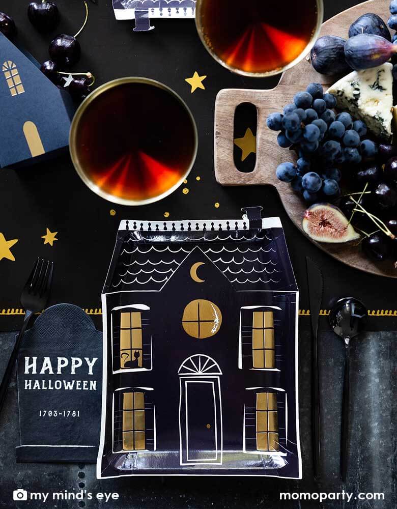 A black and gold Halloween party table featuring Momopartys's 8" x 10.5" haunted village haunted house shape plates and black tombstone shaped napkins with Happy Halloween words on them. Around the tableware, there are red drinks and black themed charcuterie board on Momopartys's mystical dark night table runner, gives a spooky vibe to an elegant Halloween tablescape.