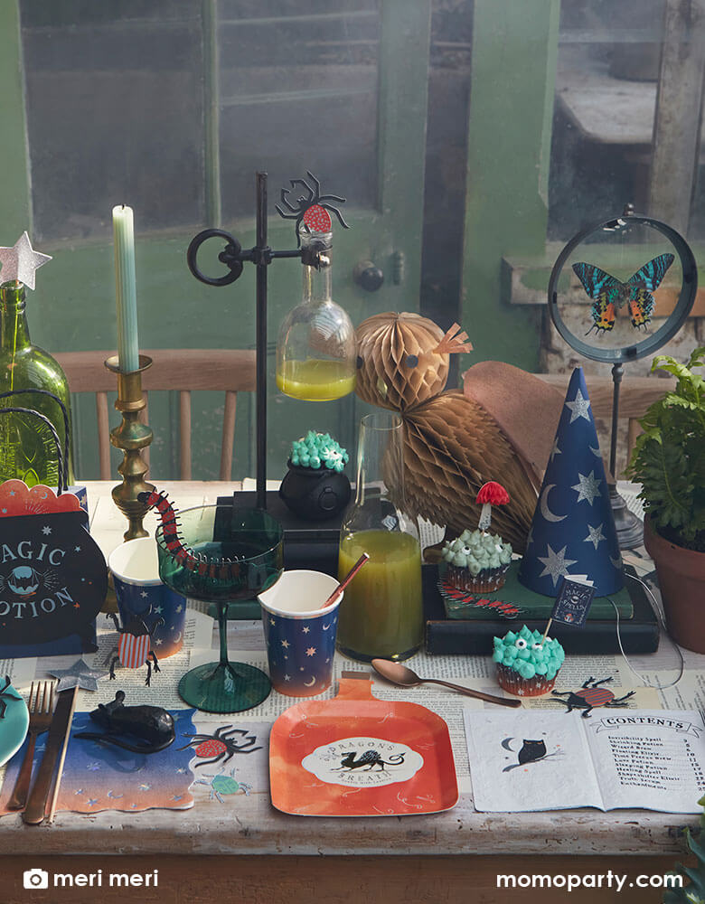 A witch and wizard party table set up featuring Momopartys's making magic party supplies collection by Meri Meri including potion bottle shaped plates, wizard spell book napkins, witch cauldron shaped party bags, owl honeycomb decoration, night ombre napkins and cups with moon and stars on them, and bug confetti scattered on the table, makes it a great inspiration for kid's harry potter themed party or a Halloween witch and wizard themed bash.