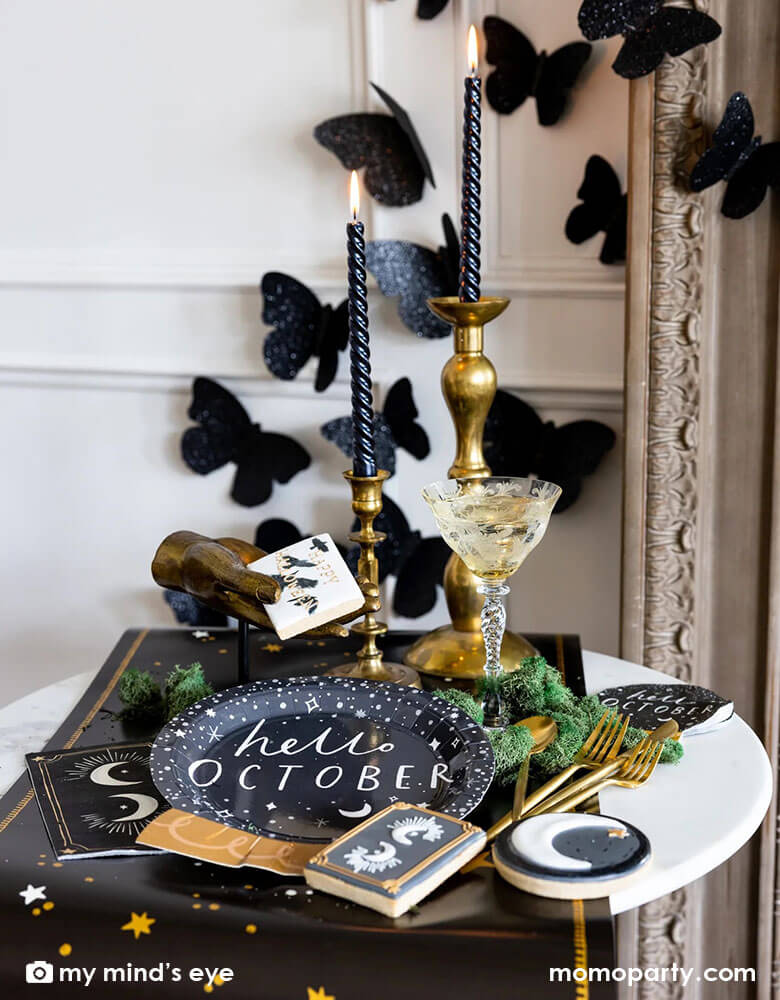 A mystical Halloween table decorated with Momopartys's moons and stars themed party supplies including a crystal ball shaped plate, moons and stars gold foil napkins, moons and stars table runner by My Mind's Eye. With gold metal candle stands holding black candles and vintage gold utensils with Halloween themed sugar cookies and black glittered butterfly decorations on the wall, all makes a perfect inspo for an elegant and chic wizard and witch themed Halloween party decorating idea.