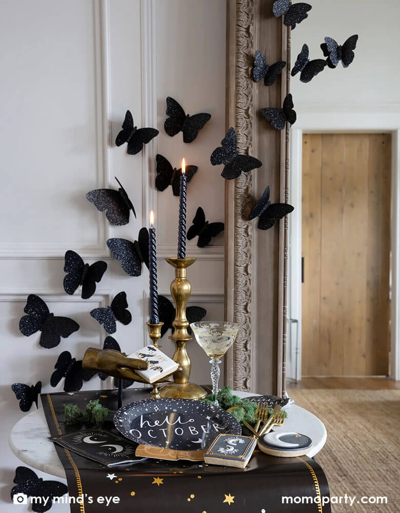 A mystical Halloween table decorated with Momopartys's moons and stars themed party supplies including a crystal ball shaped plate, moons and stars gold foil napkins, moons and stars table runner by My Mind's Eye. With gold metal candle stands holding black candles and vintage gold utensils with Halloween themed sugar cookies and black glittered butterfly decorations on the wall, all makes a perfect inspo for an elegant and chic wizard and witch themed Halloween party decorating idea.