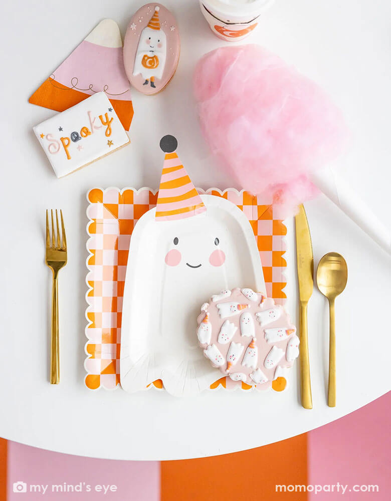 A pink Halloween party table featuring Momopartys's ghoul gang collection by My Mind's Eye including 11.5" x 6.5" ghost shaped plates, orange and pink checkered scallop edged plates and ghoul gang pink to-go cups. With coordinating decorated sugar cookies and pink cotton candy around the table, it makes an adorable pink girly Halloween party inspiration.