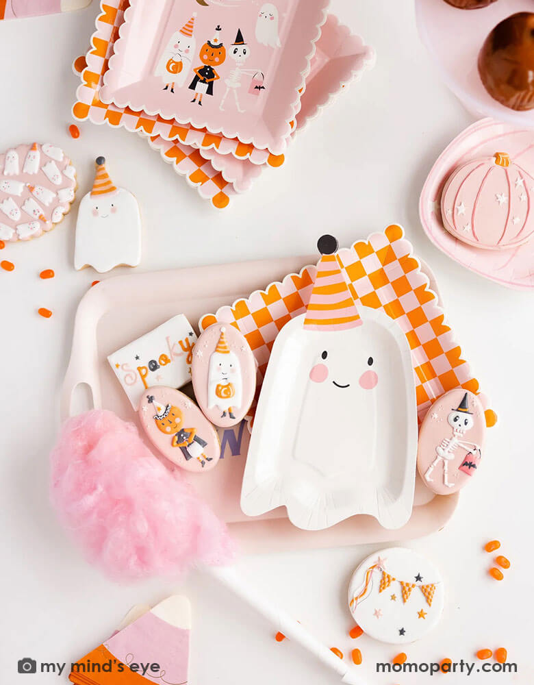 A pink Halloween party table featuring Momopartys's ghoul gang collection by My Mind's Eye including 11.5" x 6.5" ghost shaped plates, orange and pink checkered scallop edged plates, ghoul gang scene paper plates, pink pumpkin plate on a pastel pink reusable bamboo tray. With coordinating decorated sugar cookies, orange jelly beans, pink cotton candy and caramel apples around the table, it makes an adorable pink girly Halloween party inspiration.
