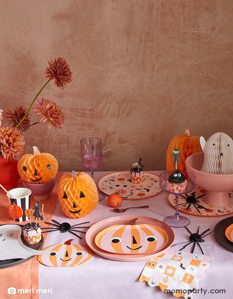 An adorable Pink Halloween party table featuring Momopartys's pink and orange striped pumpkin plates, napkins and groovy Halloween dinner plates & napkins by Meri Meri. On the table there are also cute Halloween honeycomb decorations in the shape of a ghost and pumpkin and spider with spooky cute face on them. With Halloween character cupcakes, it makes a perfect party inspiration for a cute Halloween bash for kids.