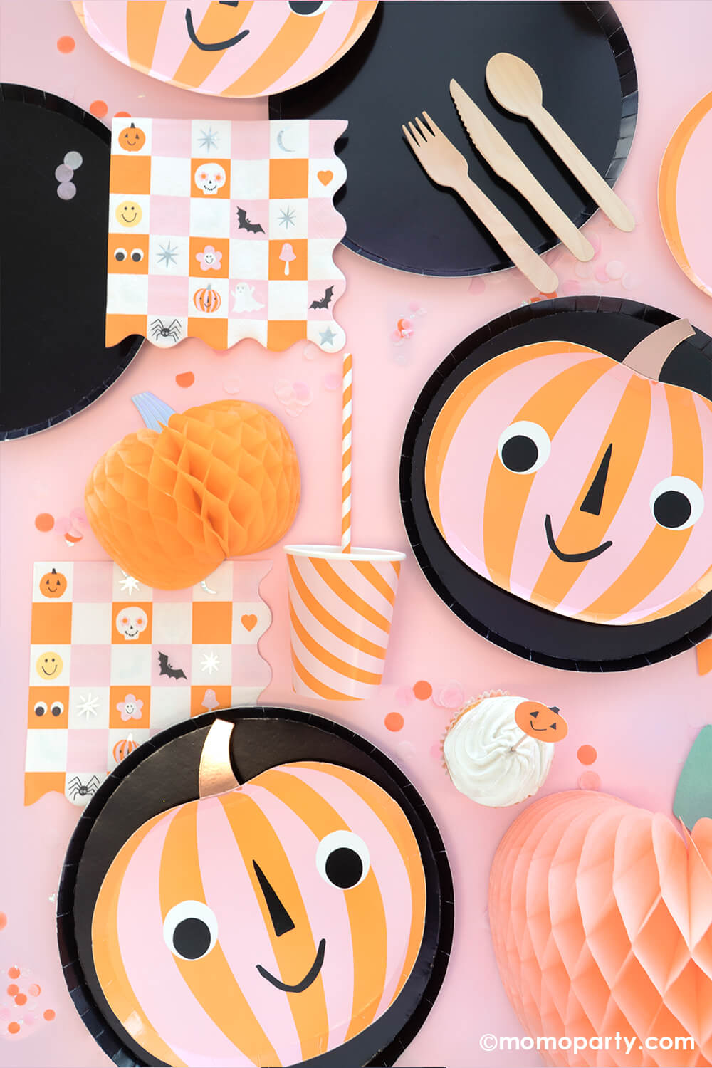 A pink girly Halloween party table featuring Momopartys's orange and pink striped pumpkin shaped plates layered on 10" black large plates, groovy Halloween checkered napkins with Halloween character designs including skulls, pumpkins, bats, spiders ghosts and googly eyes, with honeycomb pumpkins around and festive confetti in orange and pink colors, this makes a great inspiration for a not-so-scary kid-friendly Halloween party tablescape.