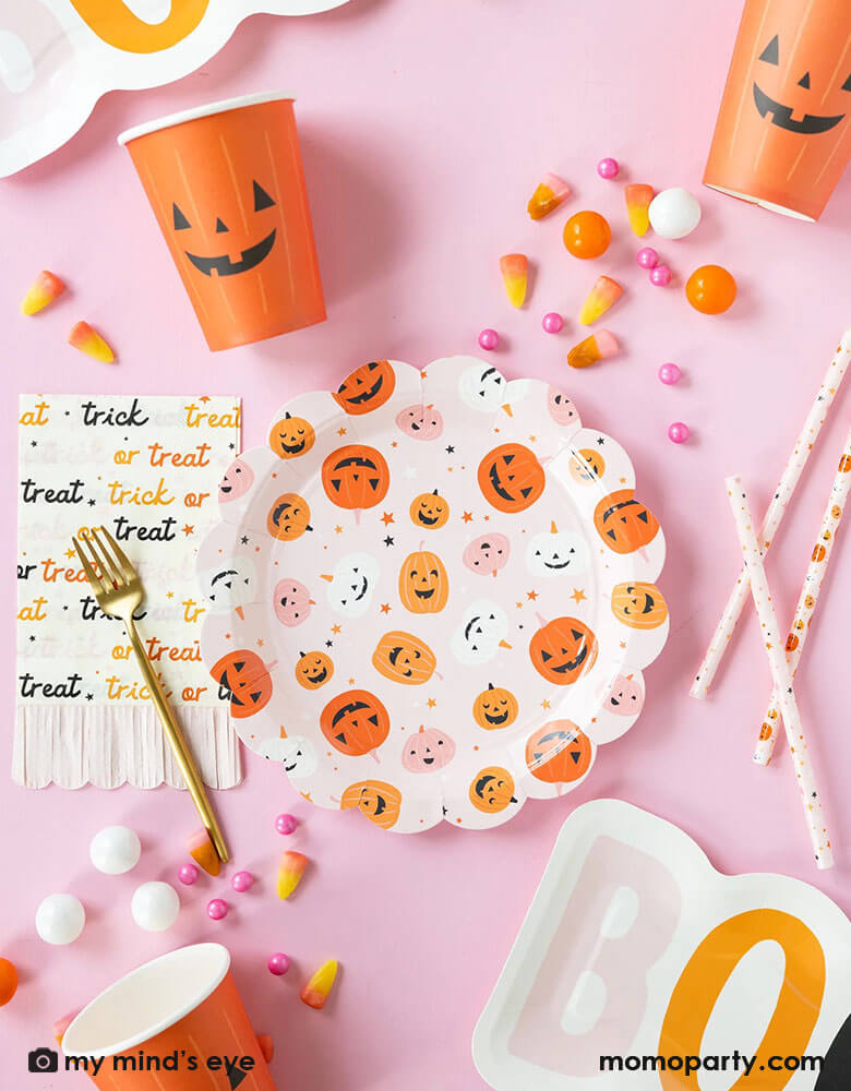 A pink Halloween party table featuring Momopartys's hey pumpkin tableware including a round pink scallop edged plates with cute jack o lantern, an orange jack o lantern party cups, pink trick or treat fringe napkins, boo shaped plates and pink pumpkin reusable straws. With pink Halloween treats like orange gum balls, candy cones, pink candies, it makes a great table setting inspiration for a cute girly kid's pink Halloween party table setting.